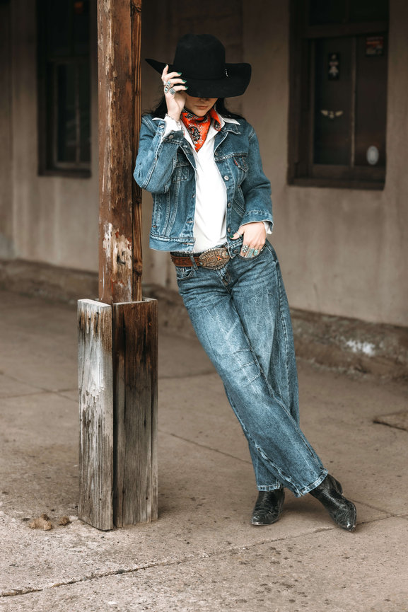 Woman in cowboy hat and denim outfit leaning against a wooden post outdoors