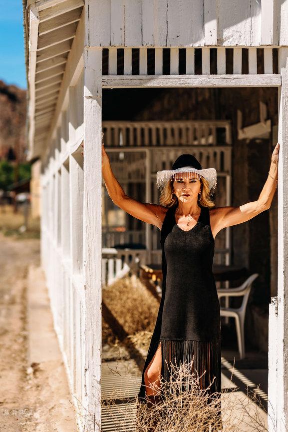 Woman in a black dress and hat posing in a rustic white doorway outdoors.