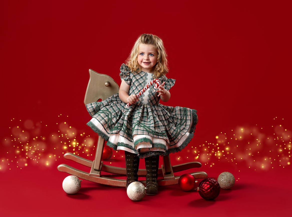 Young girl in a plaid dress sitting on a rocking horse with Christmas ornaments on a red background