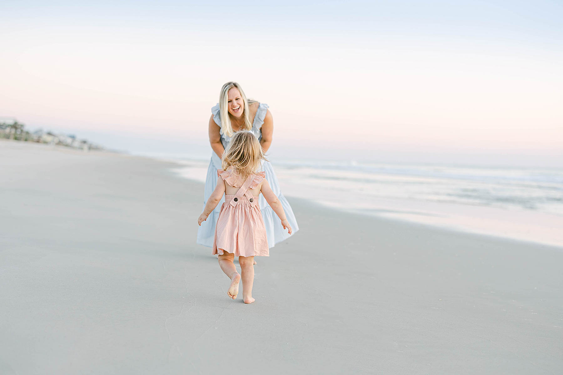 a little girl running towards her mother who is wearing a blue dress at the beach