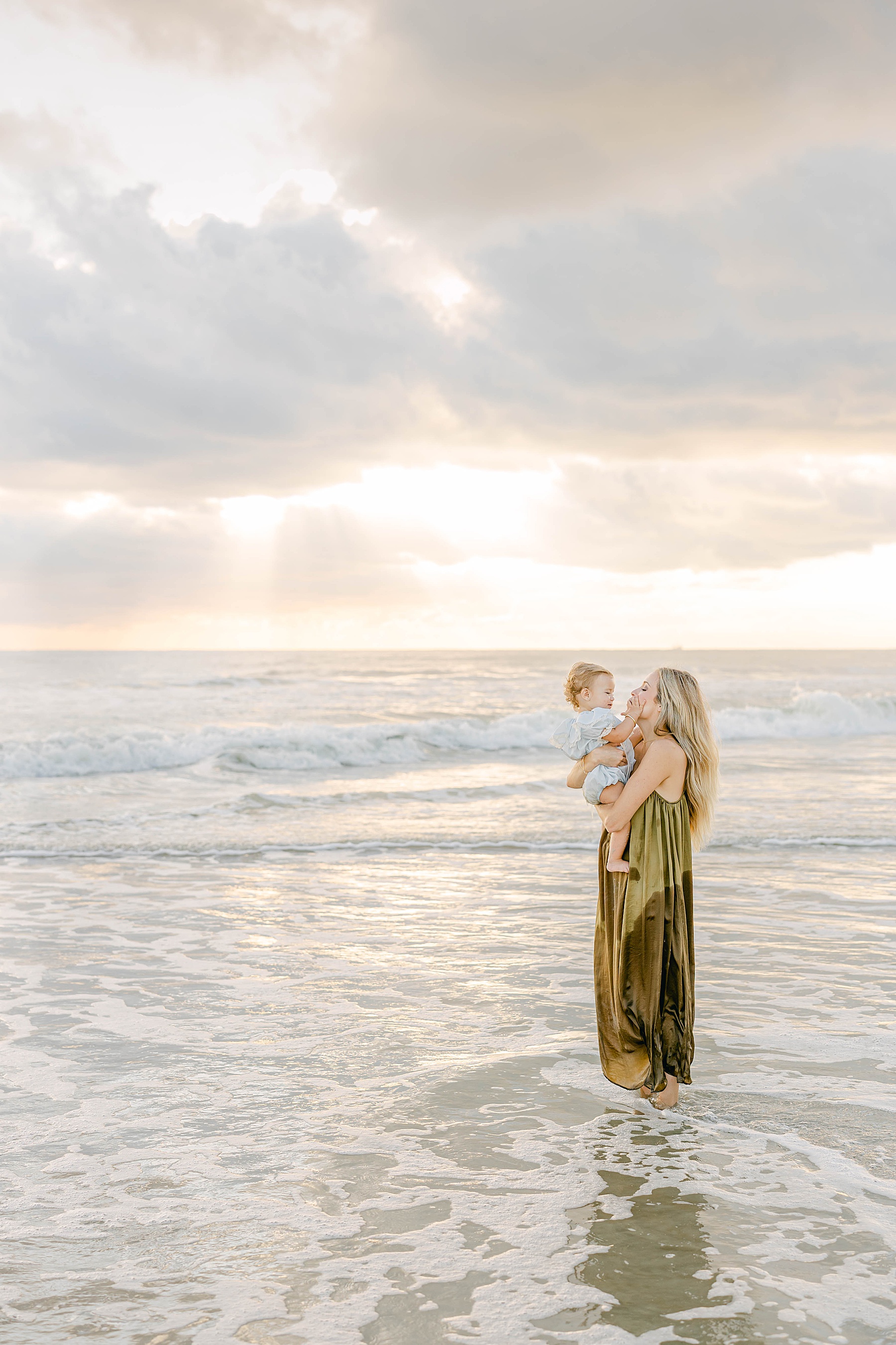 woman in green dress holding baby in blue outfit at sunrise on the beach