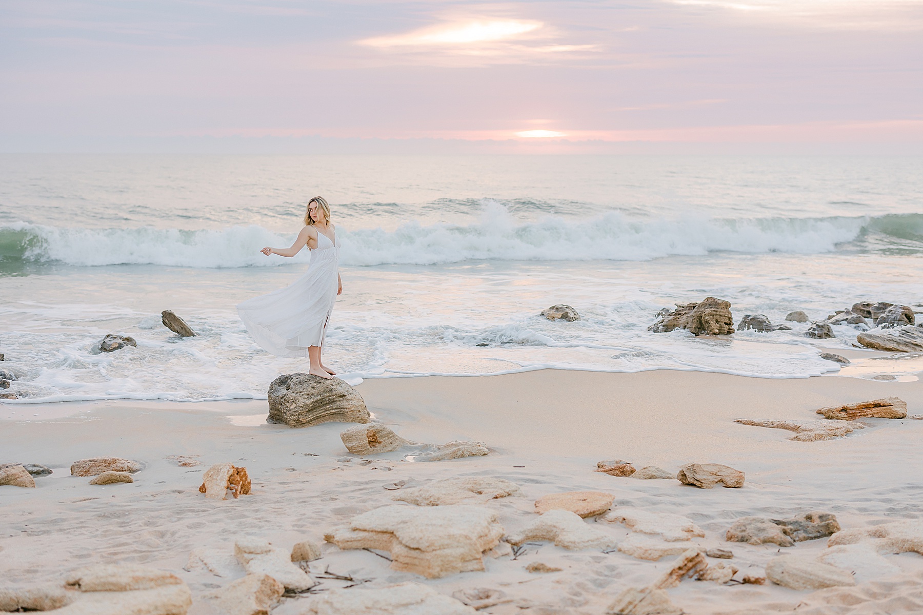 young woman standing on a rock at the beach at sunrise, light and airy pastel sunrise, light and airy photograph on the beach, st. augustine beach, white maxi dress, florida sunrise
