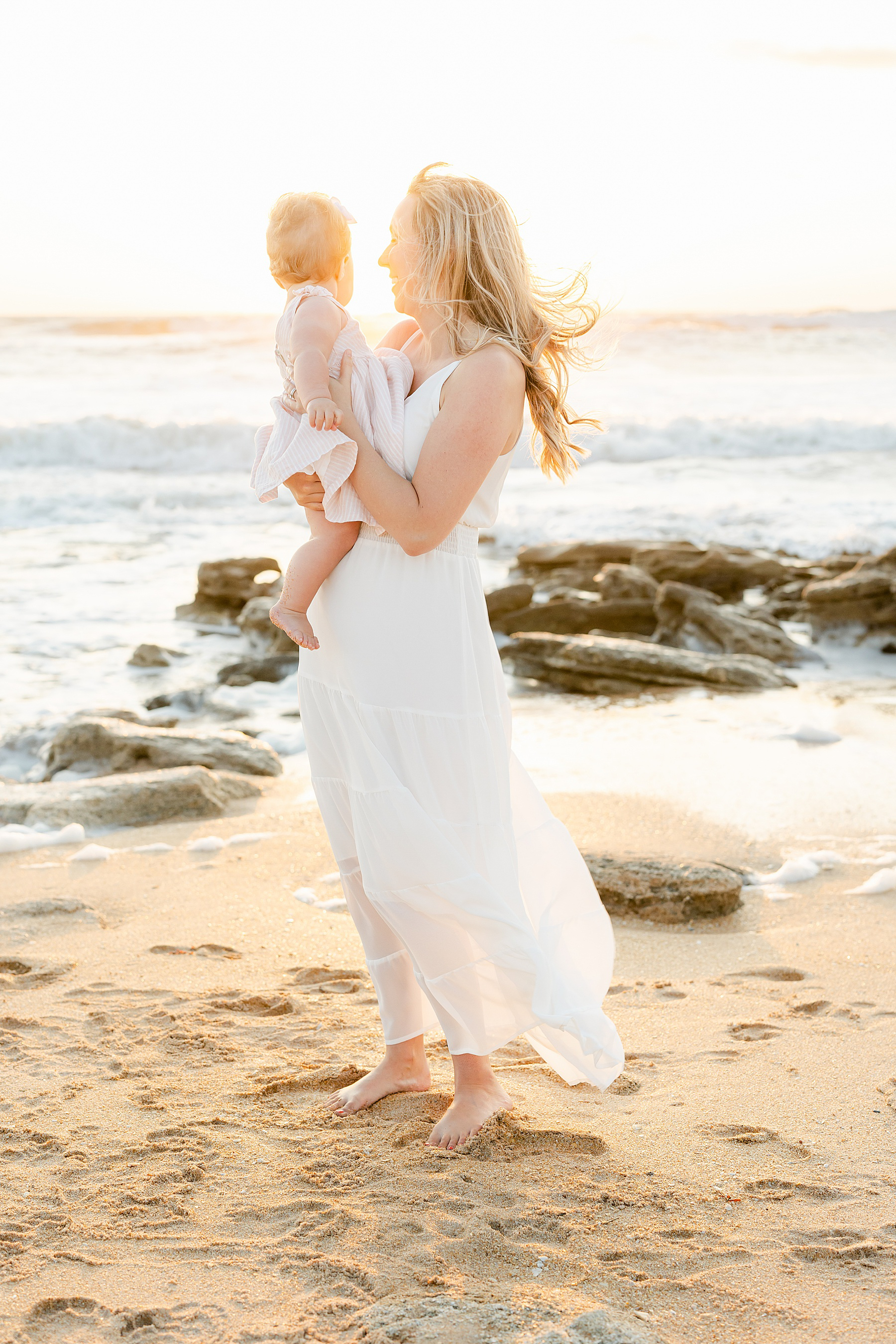 woman wearing long white dress at sunrise on the beach holding baby girl in a pink dress