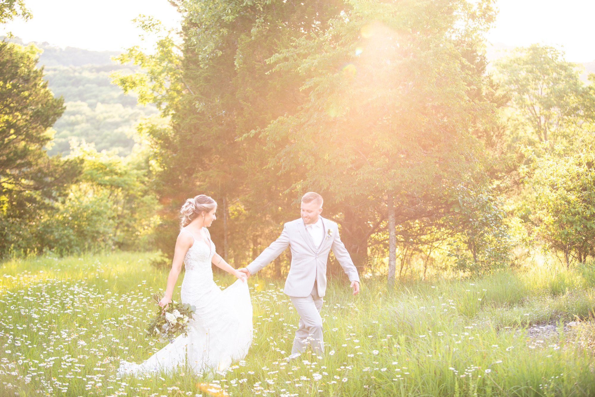 Groom leading bride by the hand in a field of flowers at sunset.