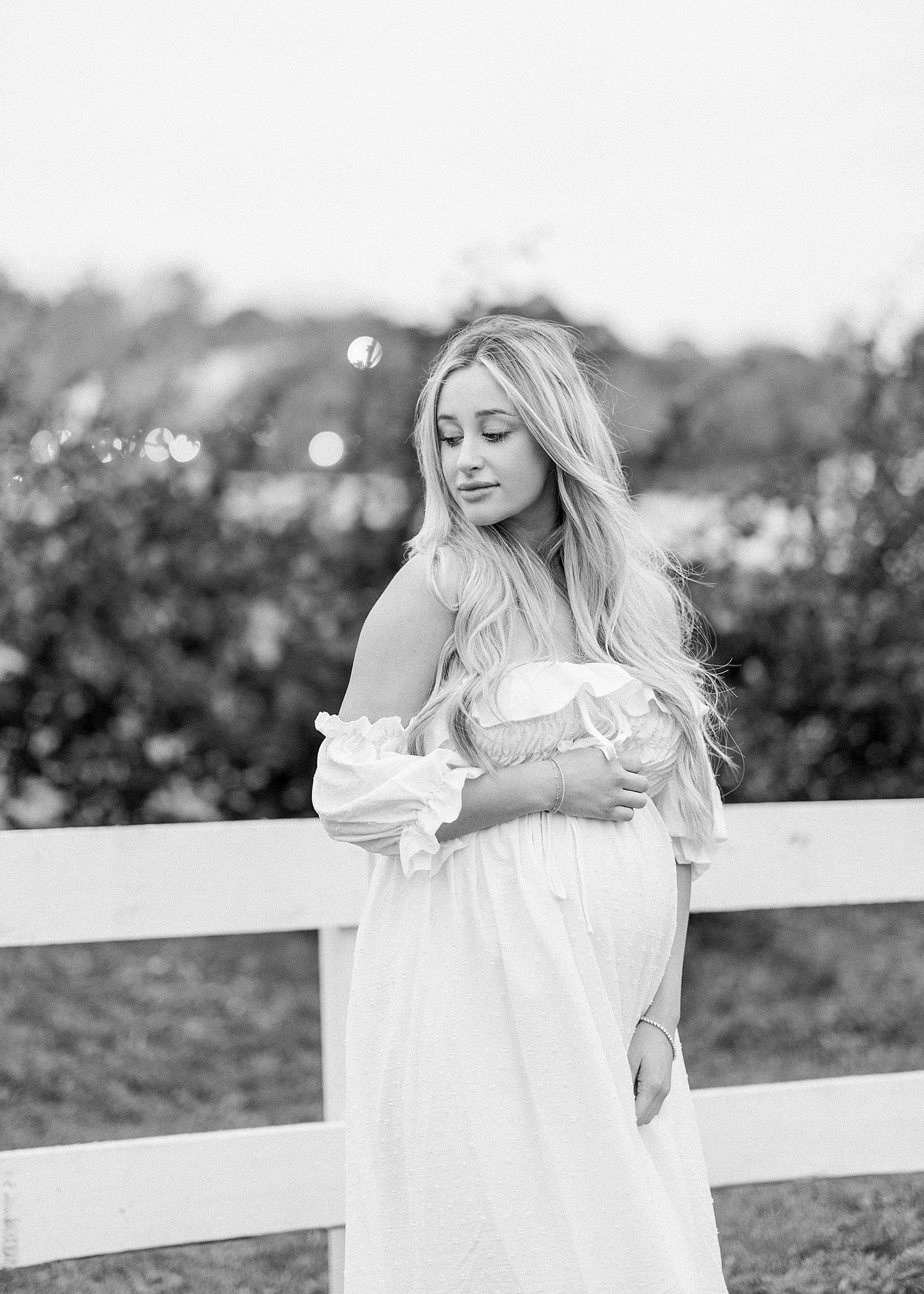 black and white maternity photo of blond haired woman in long white dress standing at Congaree and Penn Farm