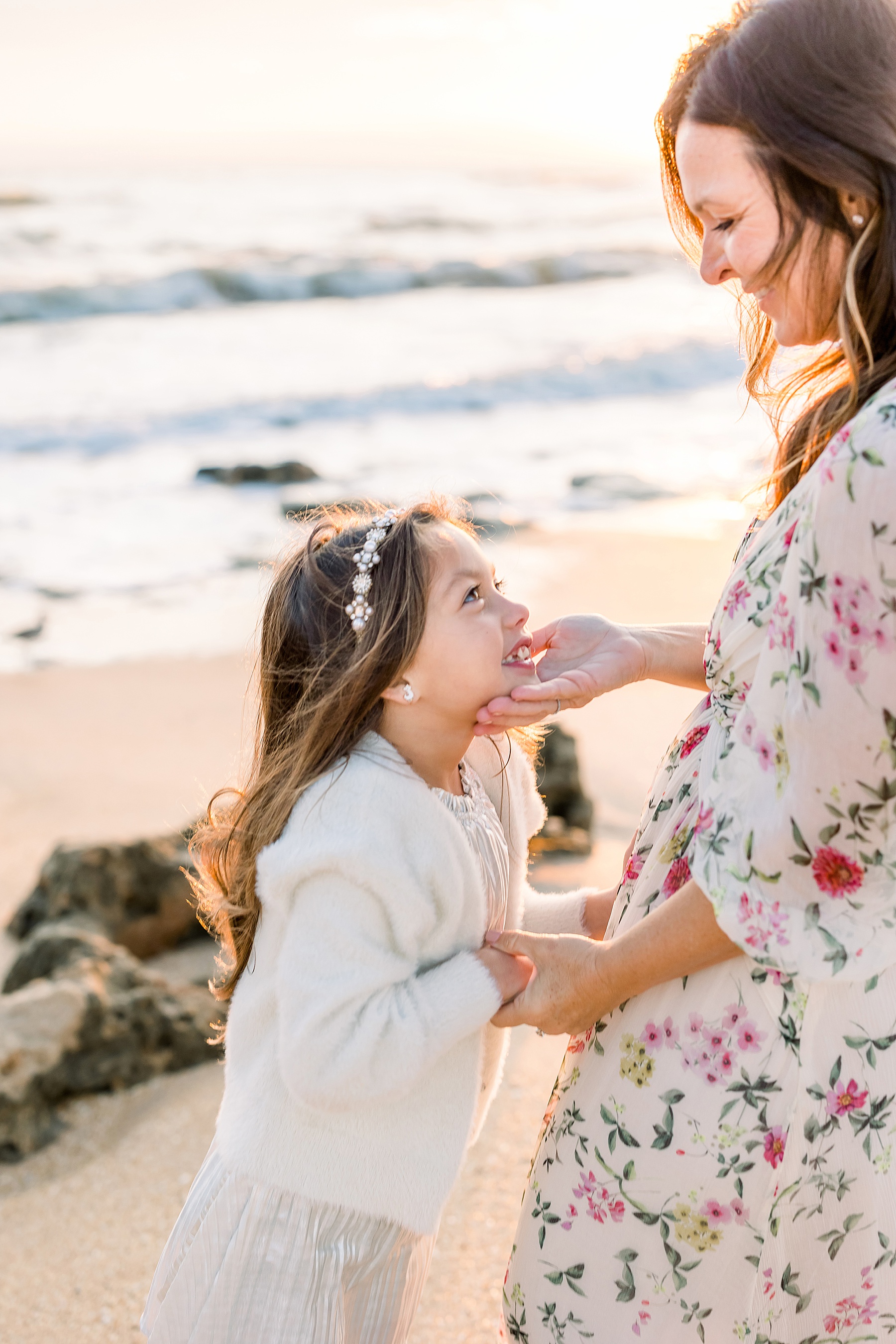 woman in floral maxi dress holding little girls chin smiling at the beach at sunrise