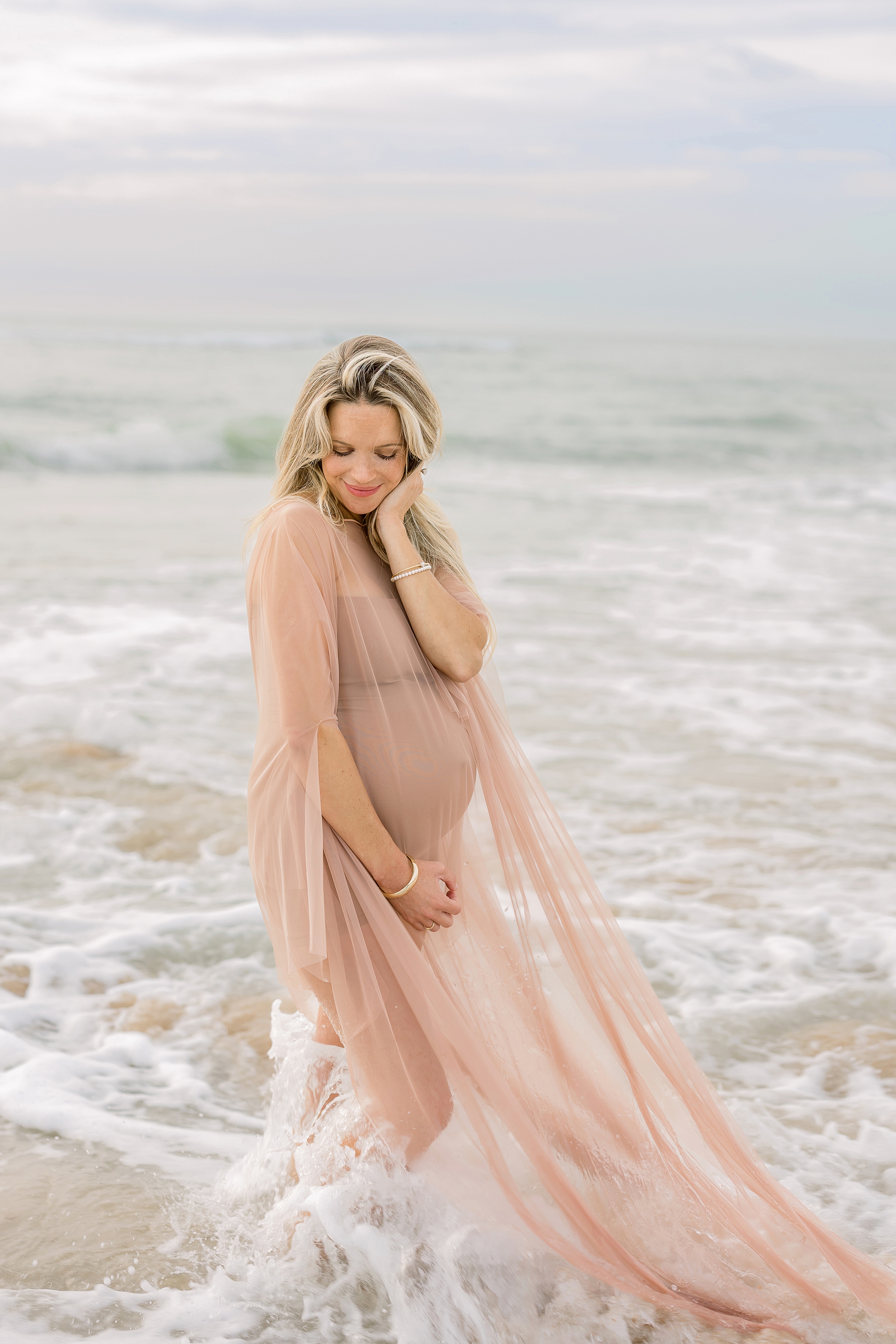 pregnant woman in airy nude sheer dress standing in the ocean on the beach