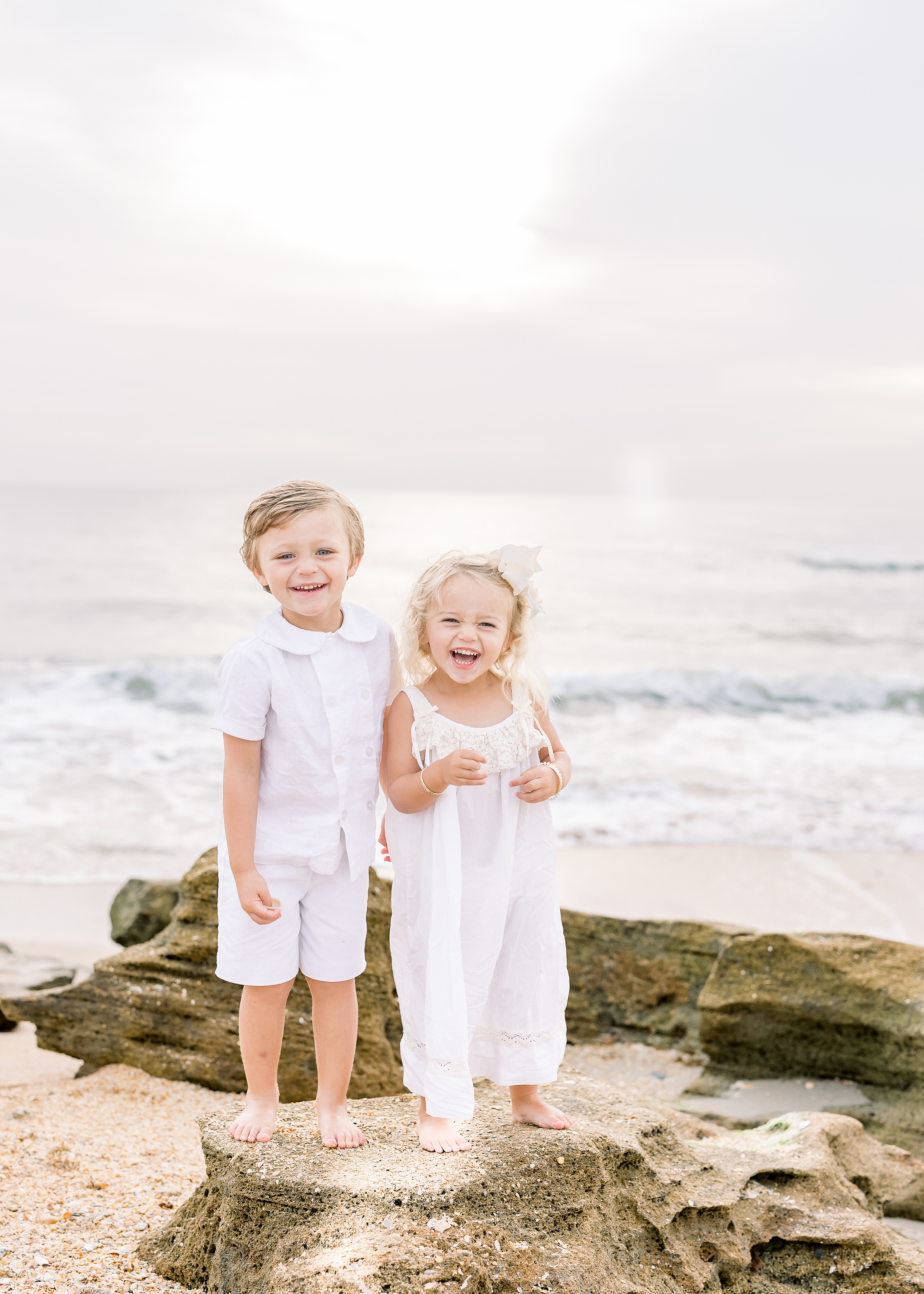little boy and little girl in white clothing standing on the rocks at the beach at sunrise