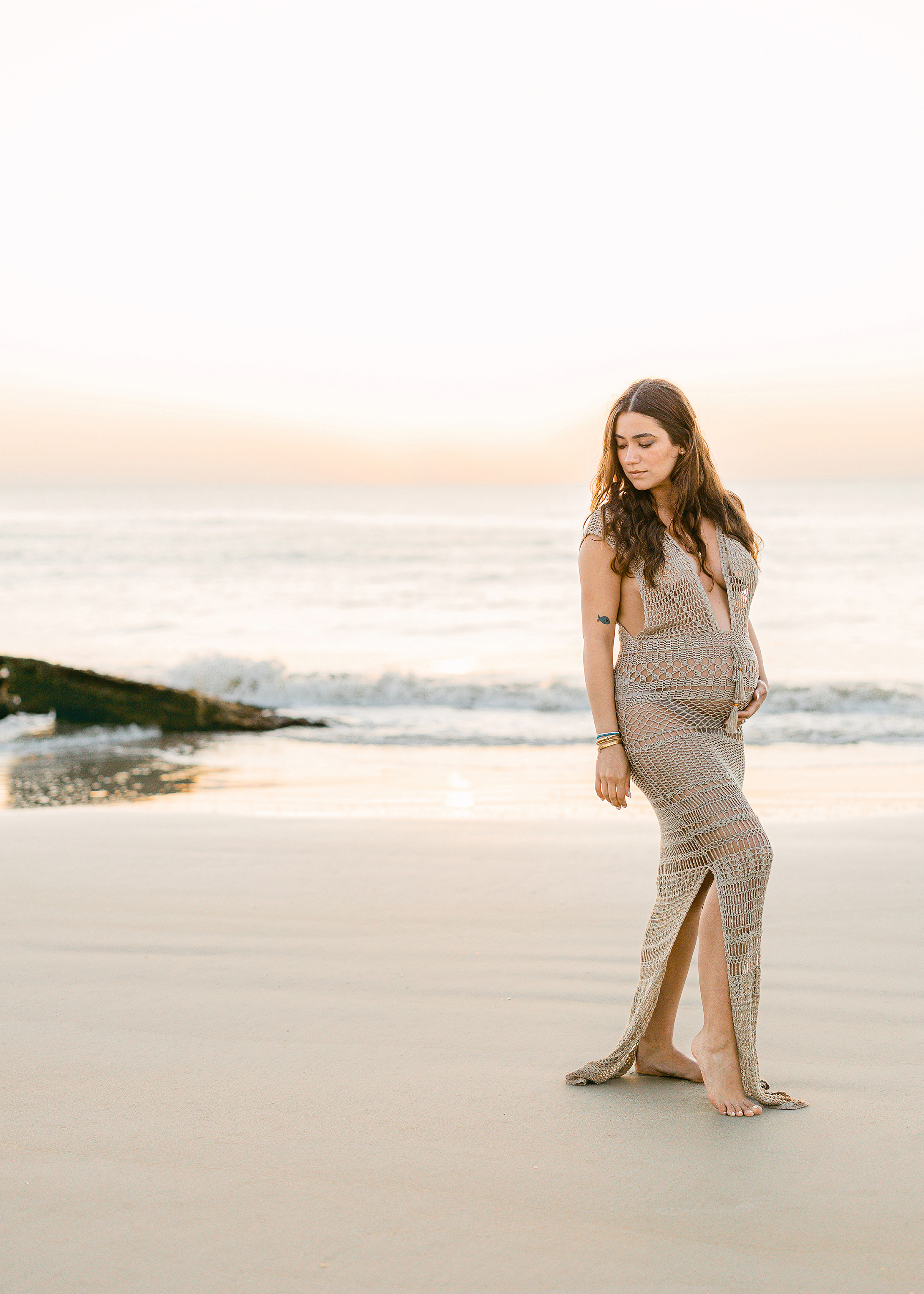 pregnant woman in shimmery fishnet maxi dress standing on the beach at sunrise