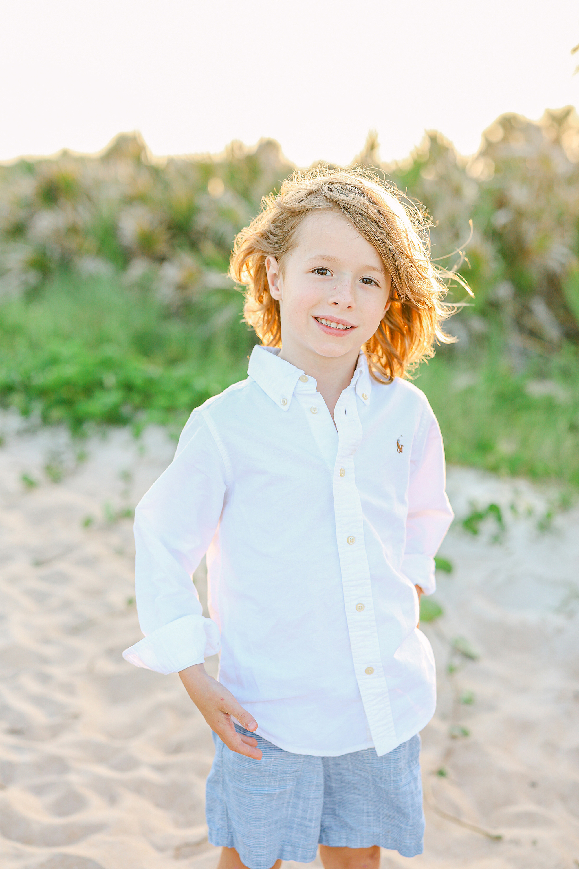 Little boy with long hair in white shirt standing on the beach at sunset.