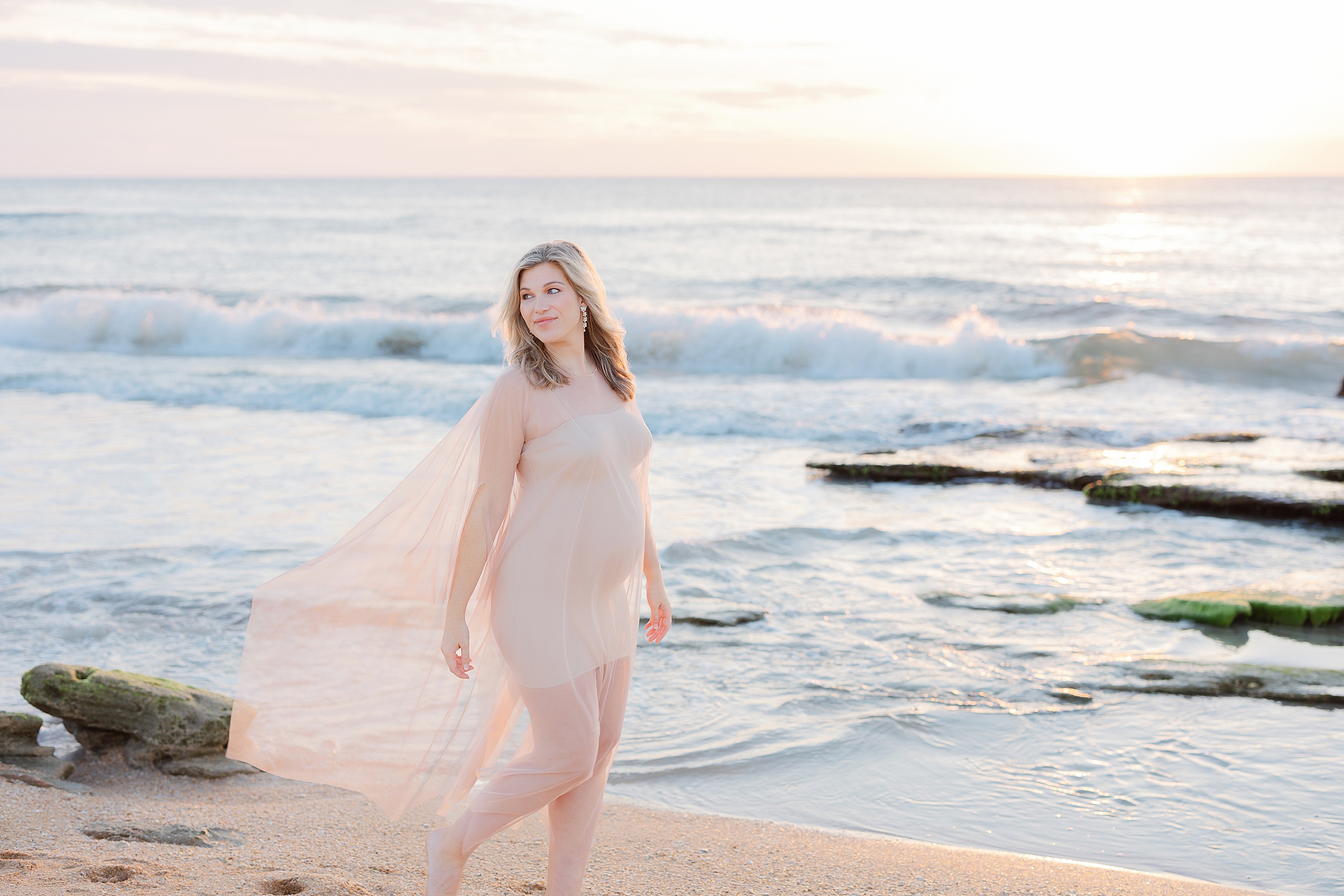 A dreamy coastal maternity portrait of a woman in nude tulle dress on the beach at sunrise.