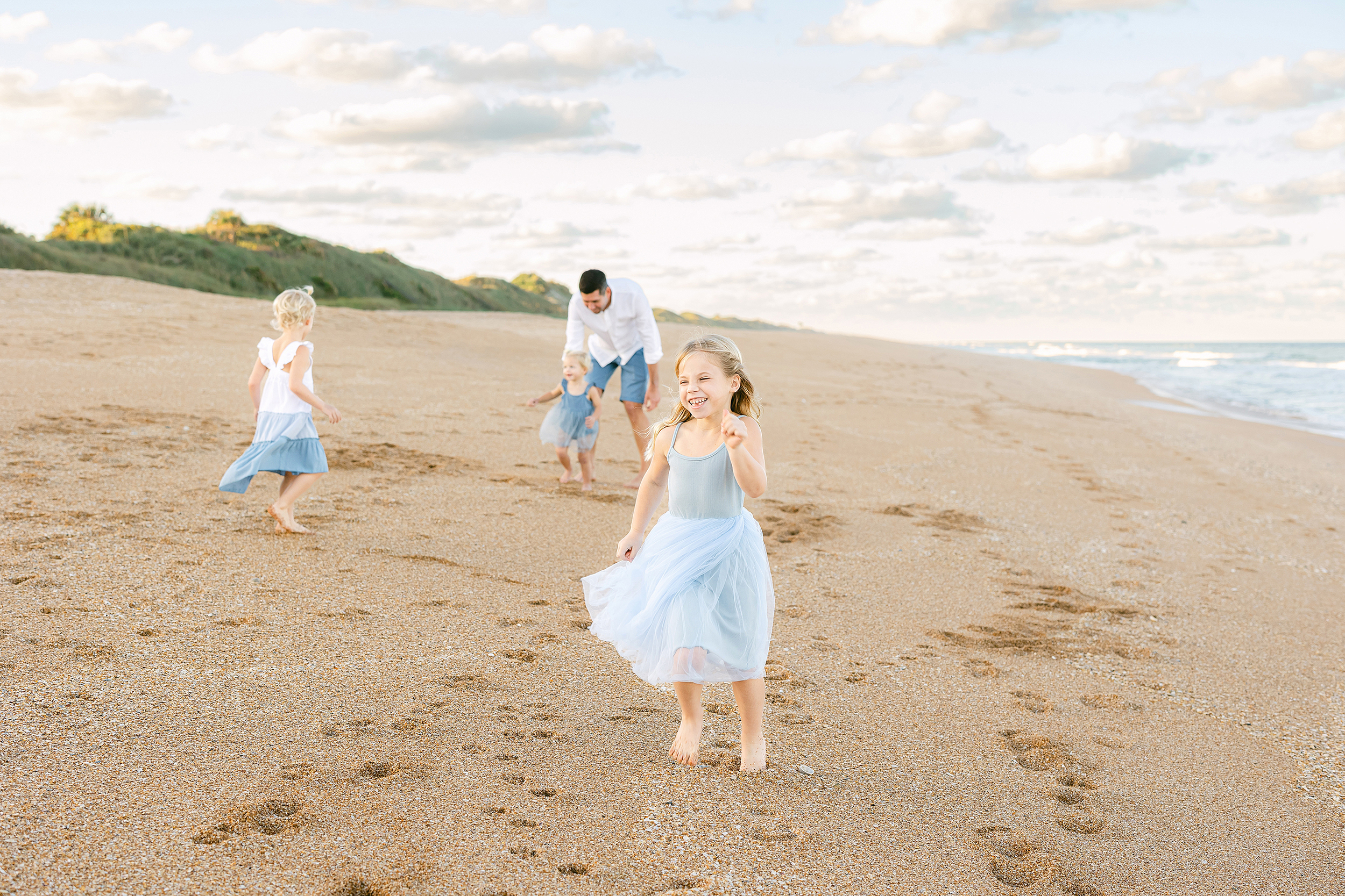 Little girl dressed in a light blue tutu dress chases her family on the beach at sunset.