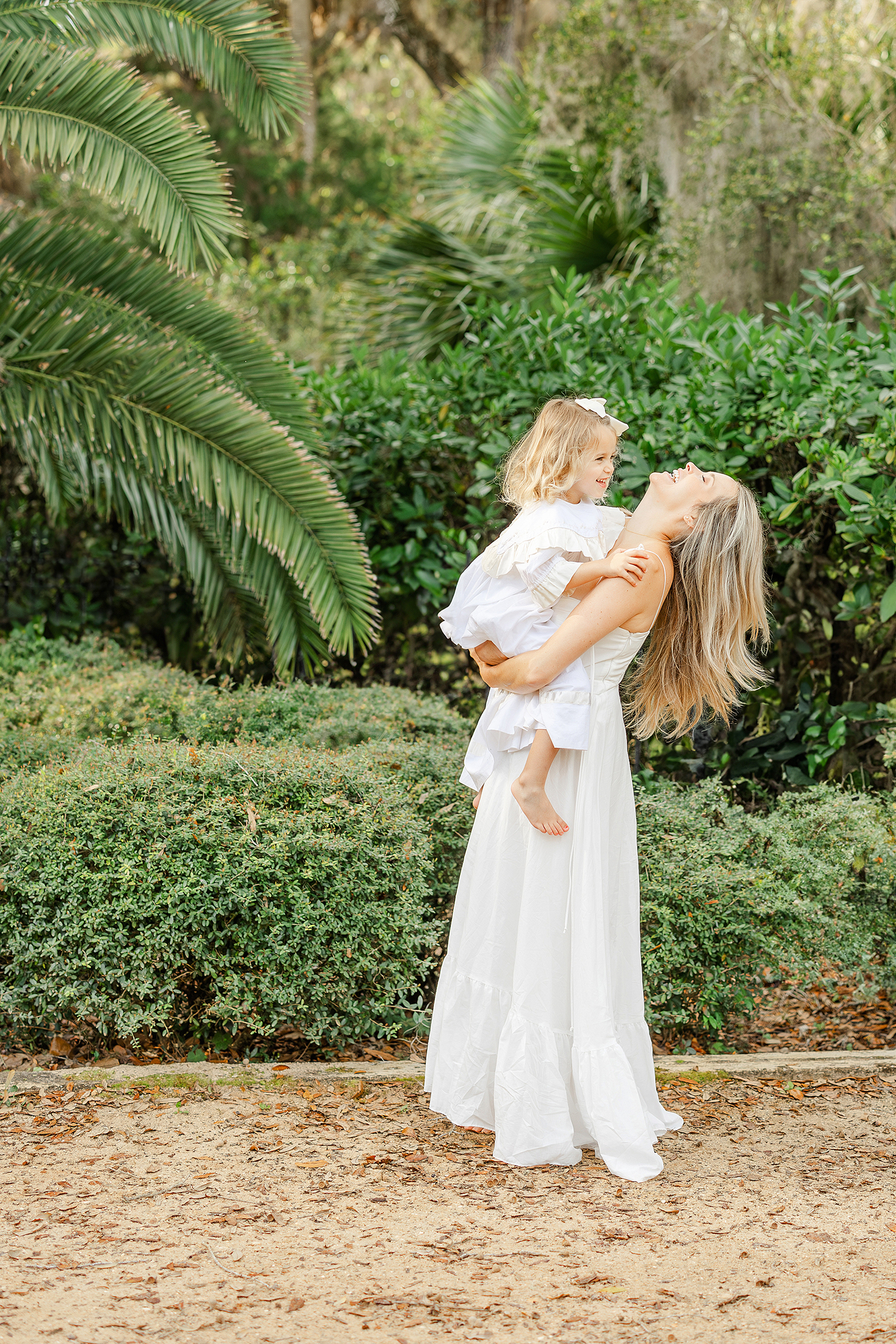 A woman in a long white dress holding a little girl in a white dress at Washington Oaks Gardens State Park.