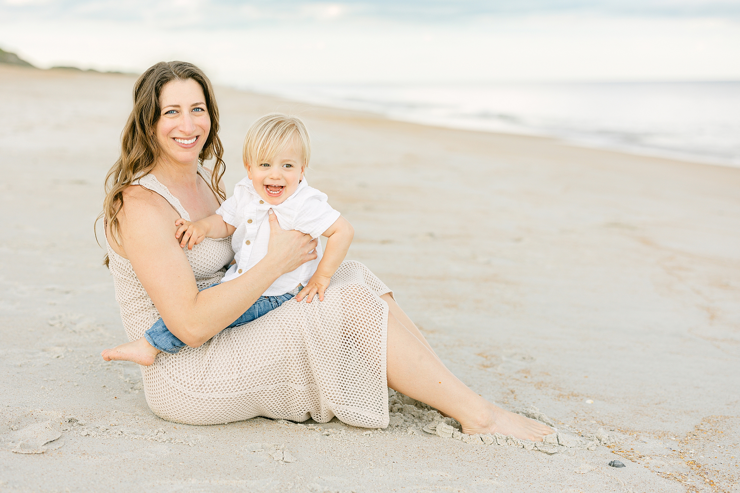 A woman in a tan crochet dress holds a little boy with blond hair on her lap on the beach.