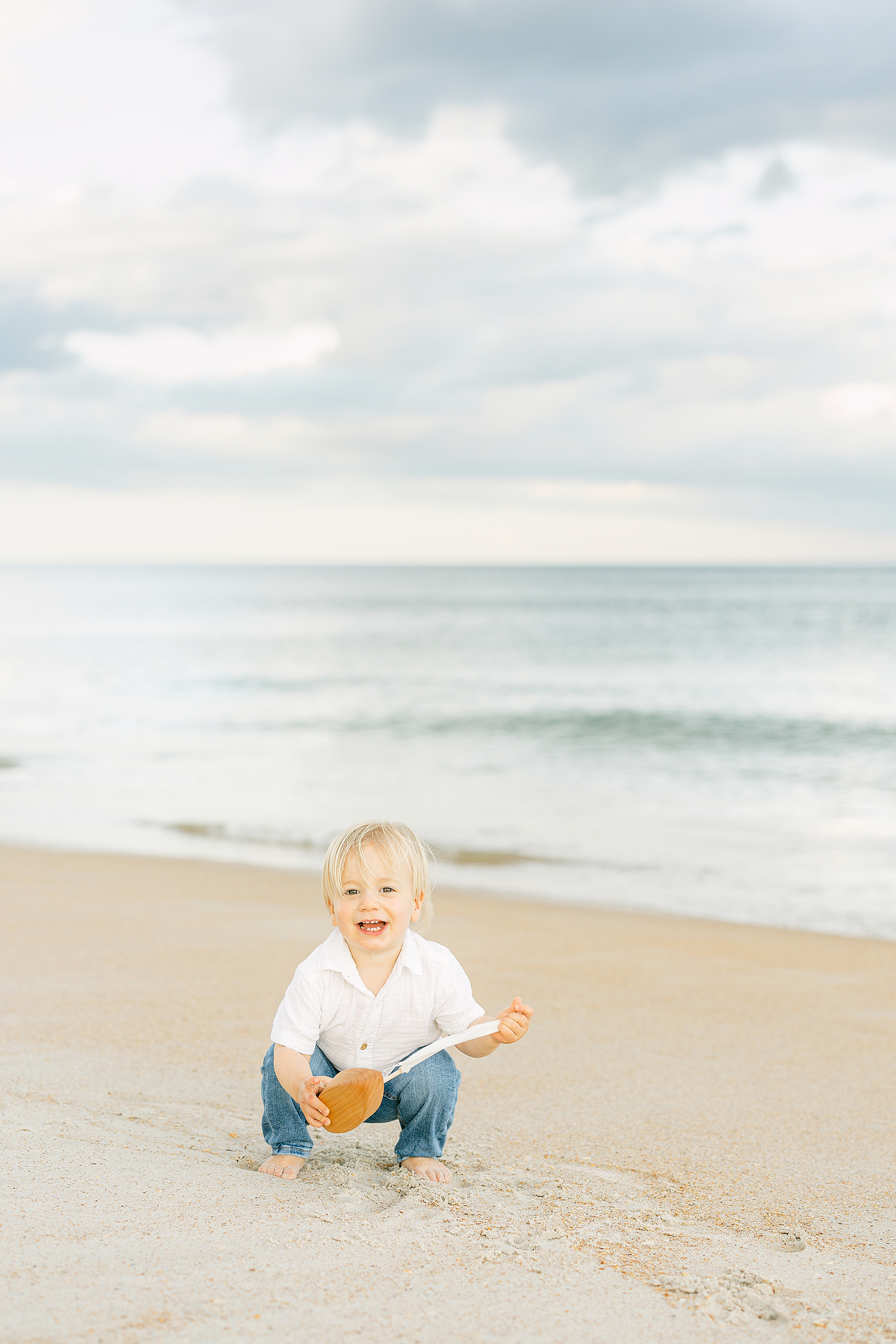 A little baby boy with blond hair wearing a white shirt and jeans holds a wooden boat on the beach.