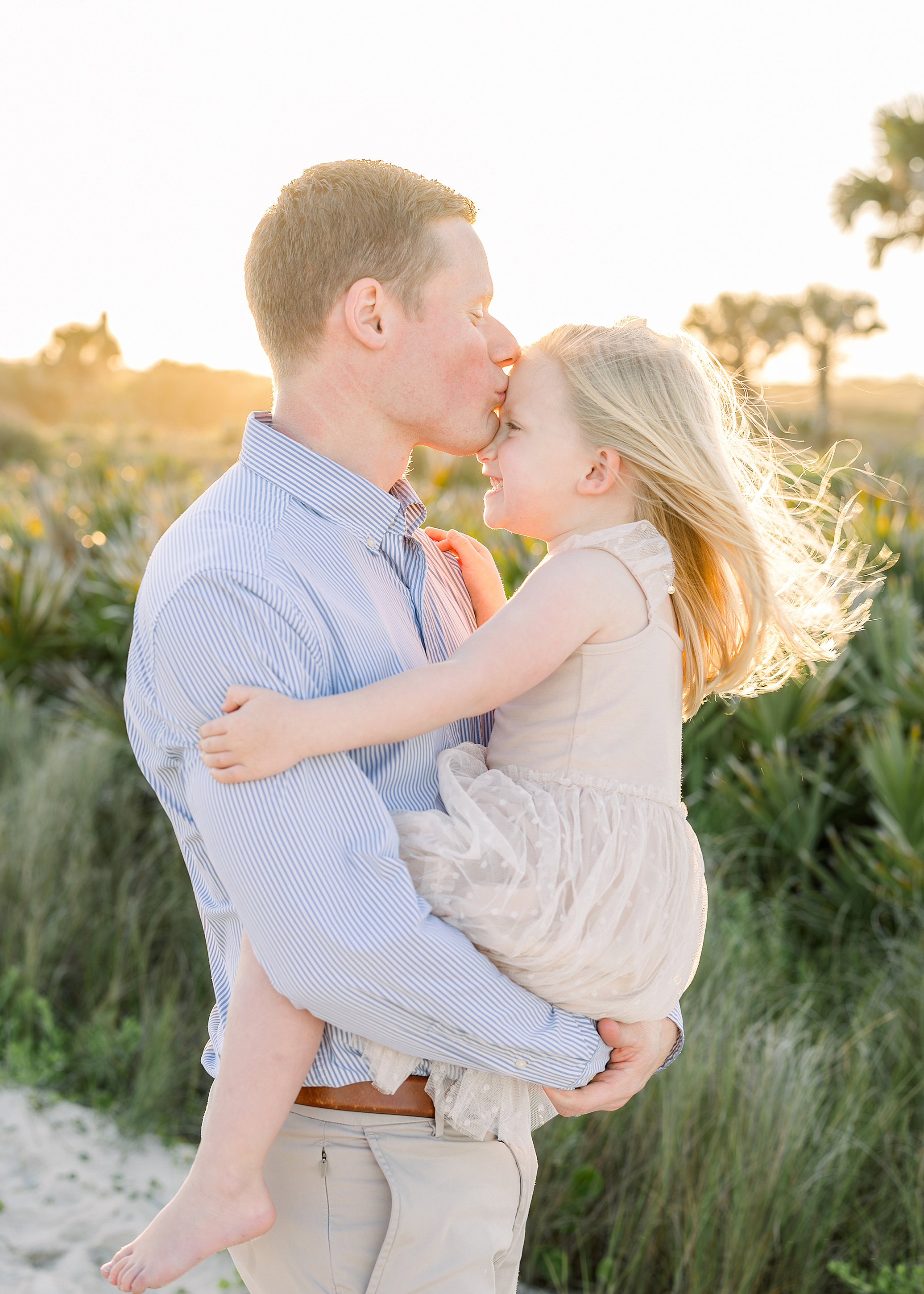 A father in a blue shirt kissing his little girl on the forehead at the beach at sunset.