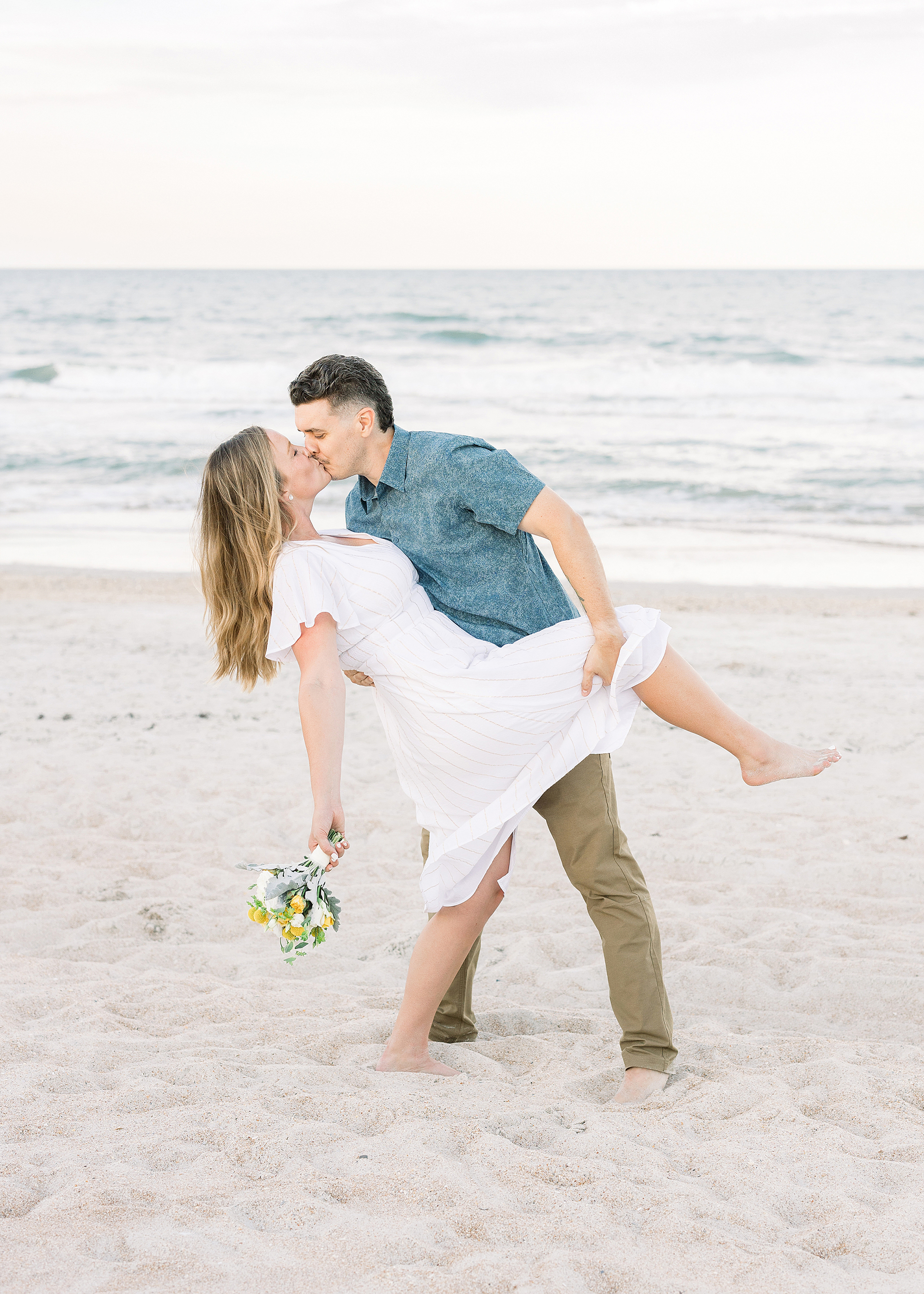 A man dips a woman in a white dress holding a bouquet back on the beach at sunset.