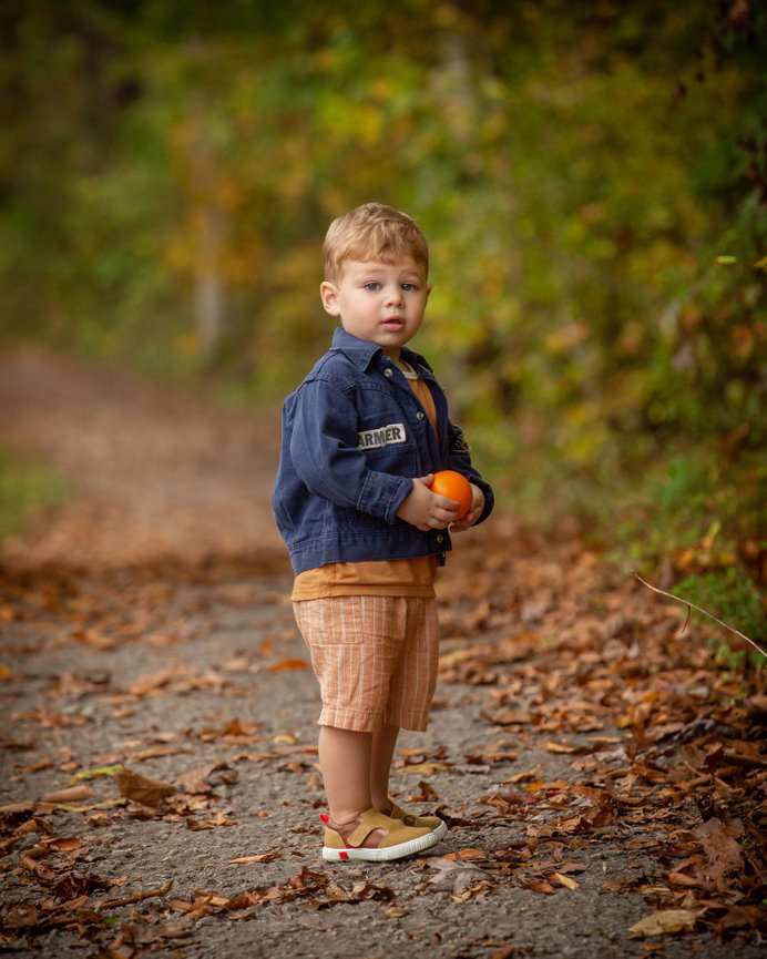 Toddler standing on a leaf-covered path, holding an orange, wearing a denim jacket and plaid shorts.