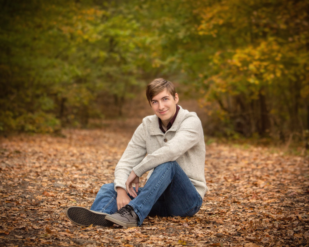 Young man sitting on a forest floor covered in autumn leaves, wearing a gray sweater and jeans, with trees in the background.