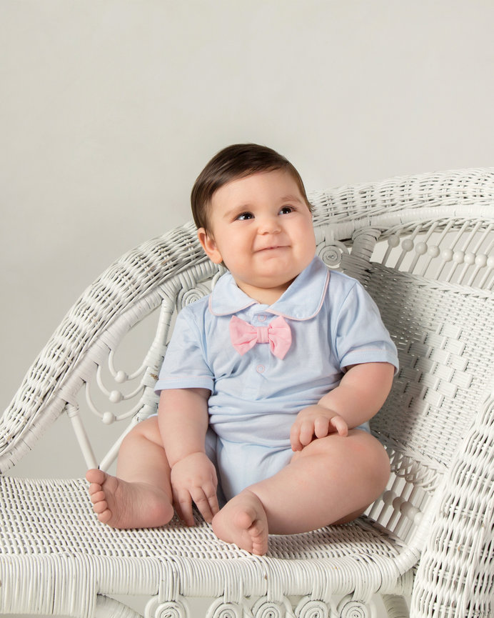 Baby in a light blue outfit with a pink bow tie sitting on a white wicker chair.