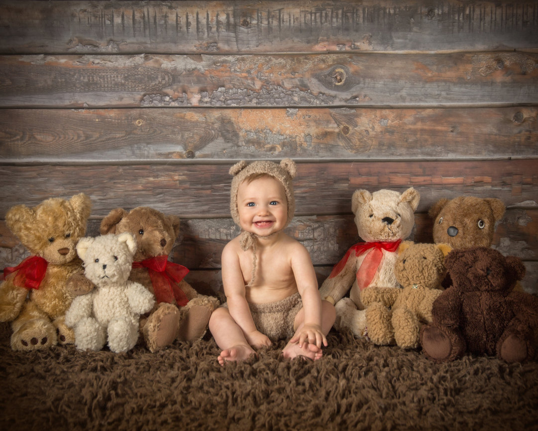 Baby in bear costume sitting on brown rug with stuffed bears in front of wooden backdrop.