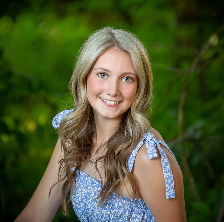 A woman with long blonde hair smiles, wearing a blue floral dress, in a green outdoor setting.