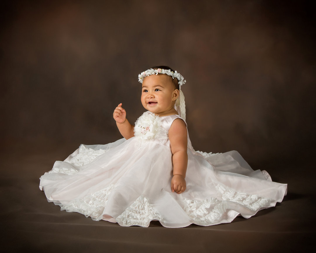Baby in a white dress and floral headband sitting on a brown background.