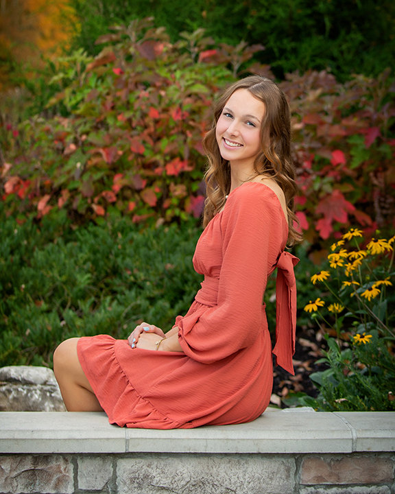 A woman in an orange dress sits on a stone ledge, surrounded by lush greenery and colorful flowers.
