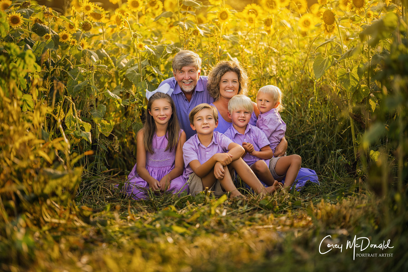Family sitting together in a sunflower field, all smiling, wearing purple and white outfits, with sunlight filtering through.