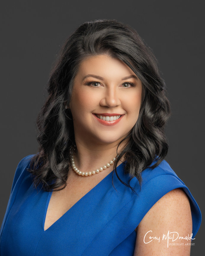 A woman with dark hair, wearing a blue dress and pearl necklace, smiles against a gray background.