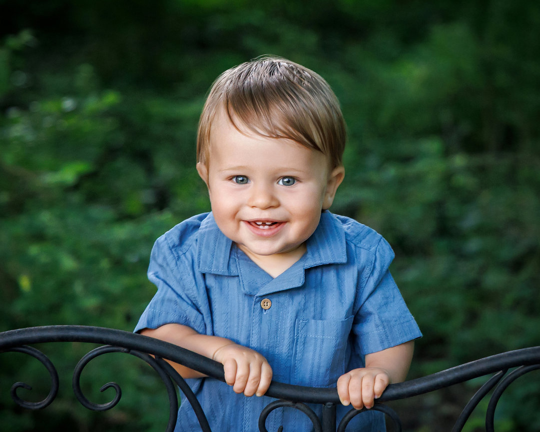 Smiling toddler in a blue shirt standing by a black wrought iron fence with greenery in the background.