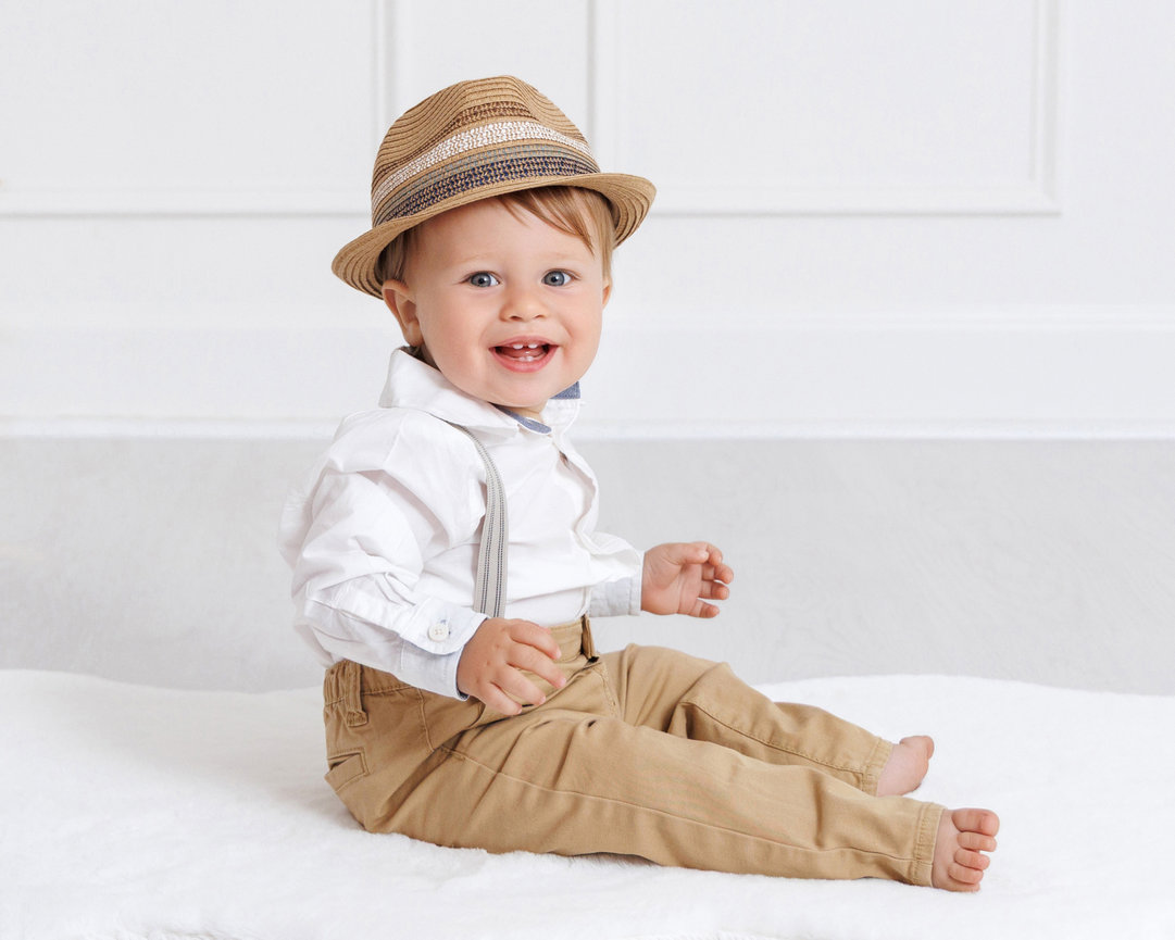 Smiling toddler in a hat wearing a white shirt with suspenders and beige pants, sitting on a soft white surface.