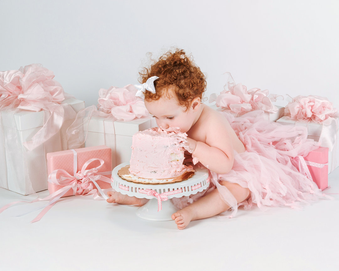 Curly-haired toddler in pink tutu eagerly eating a pink cake surrounded by wrapped gifts.