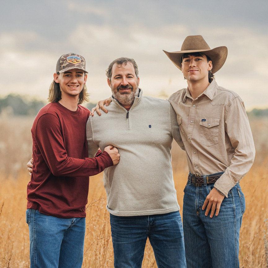 A bearded man stands outdoors between two younger men, one in a cowboy hat, with a field and cloudy sky in the background.