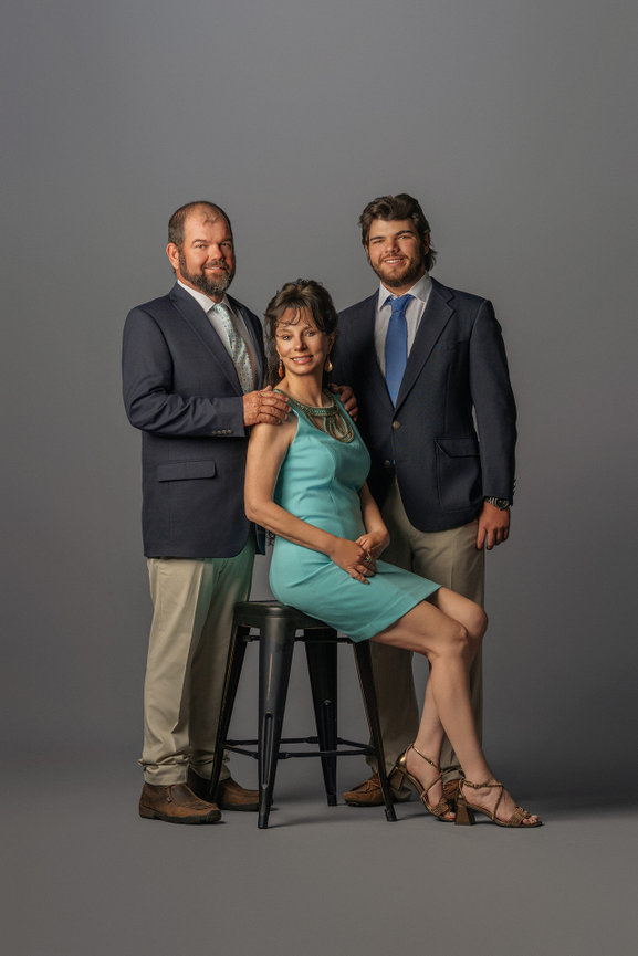 A seated woman in a turquoise dress, flanked by two men in suits against a neutral backdrop.