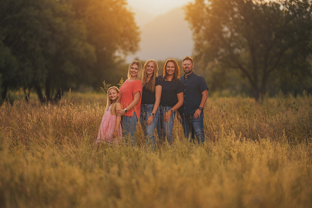 A family of five stands together in a sunlit field with trees in the background.