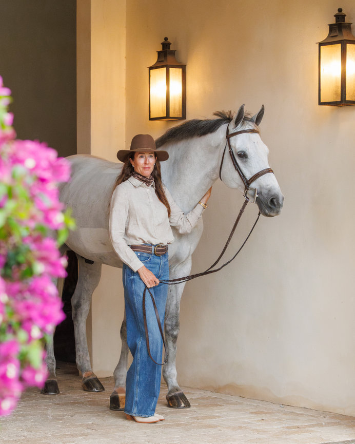 Cowgirl standing beside a white horse in a stable corridor with warm lantern light.