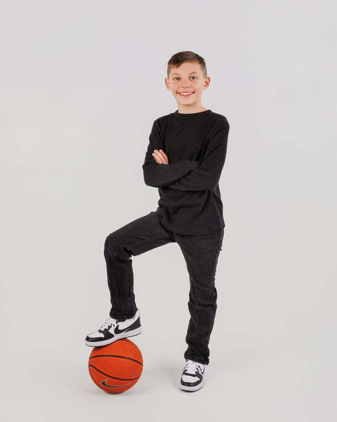 Smiling boy in black clothes posing with one foot on a basketball against a white background