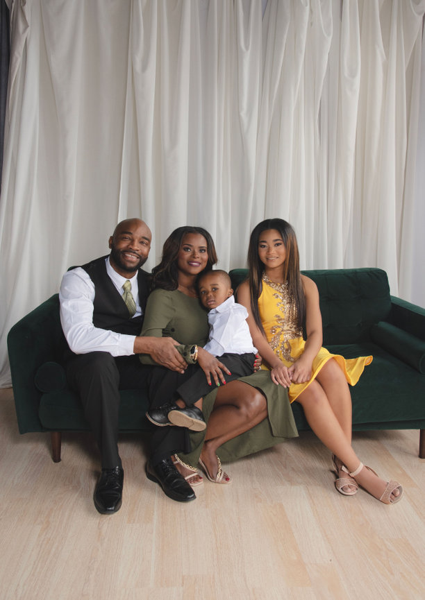 Family portrait of four, seated together on a green couch, smiling warmly against a soft, draped backdrop.