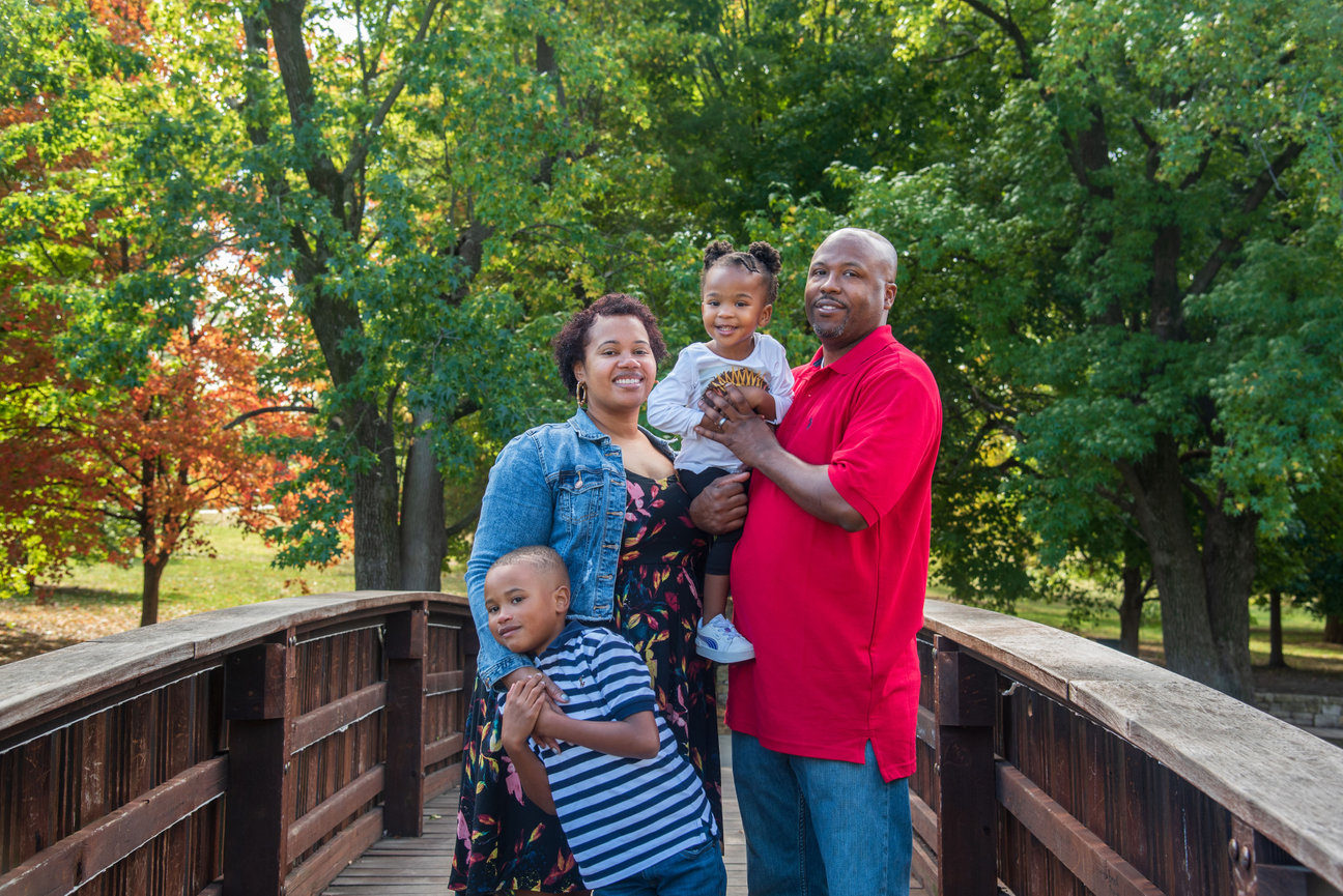 A happy family of four poses together on a bridge surrounded by lush greenery.