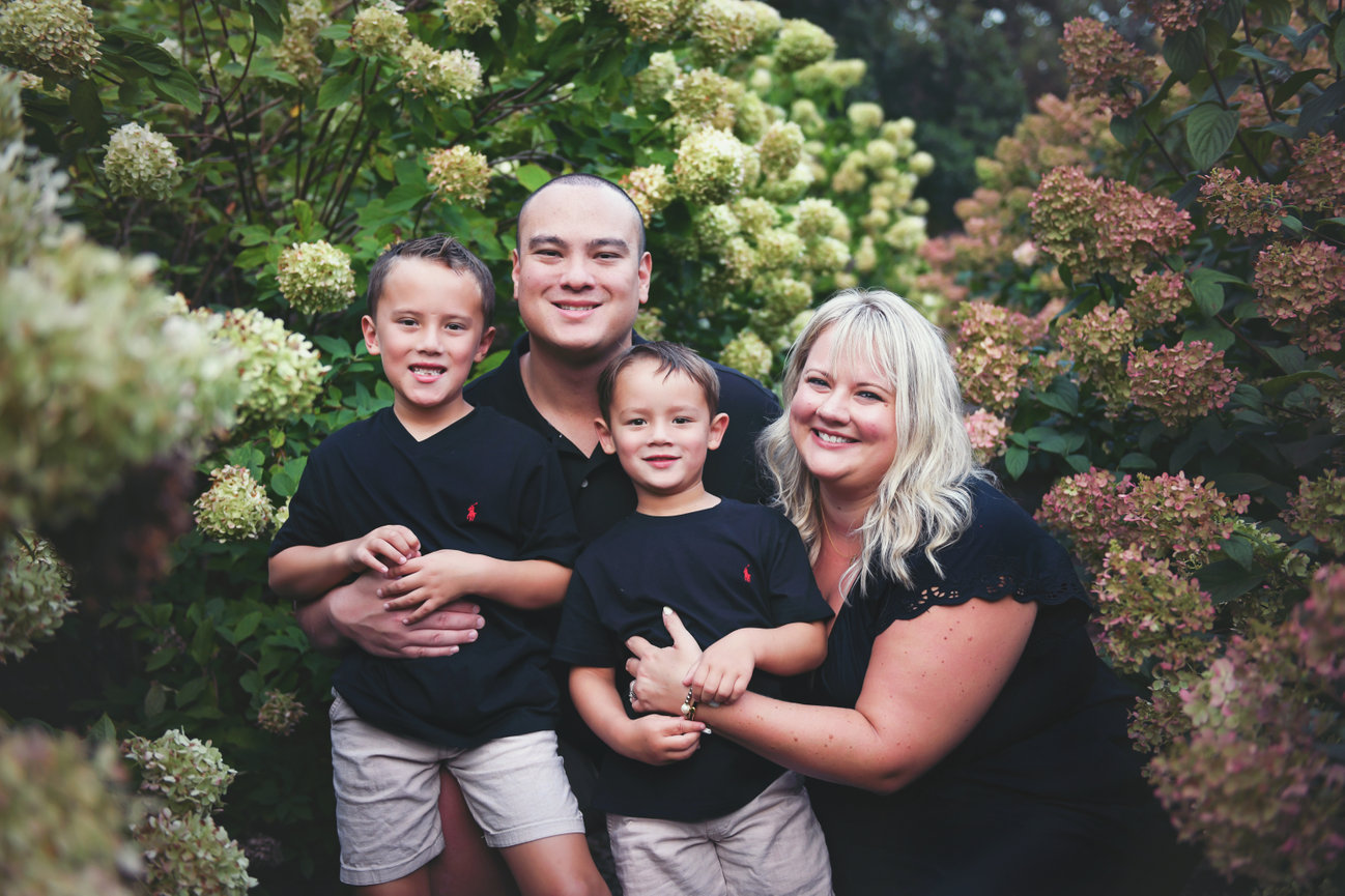 Smiling family of four in black shirts and khaki shorts surrounded by blooming hydrangeas during a vibrant summer session in Clemmons, NC at Tanglewood park by Megan Gioeli