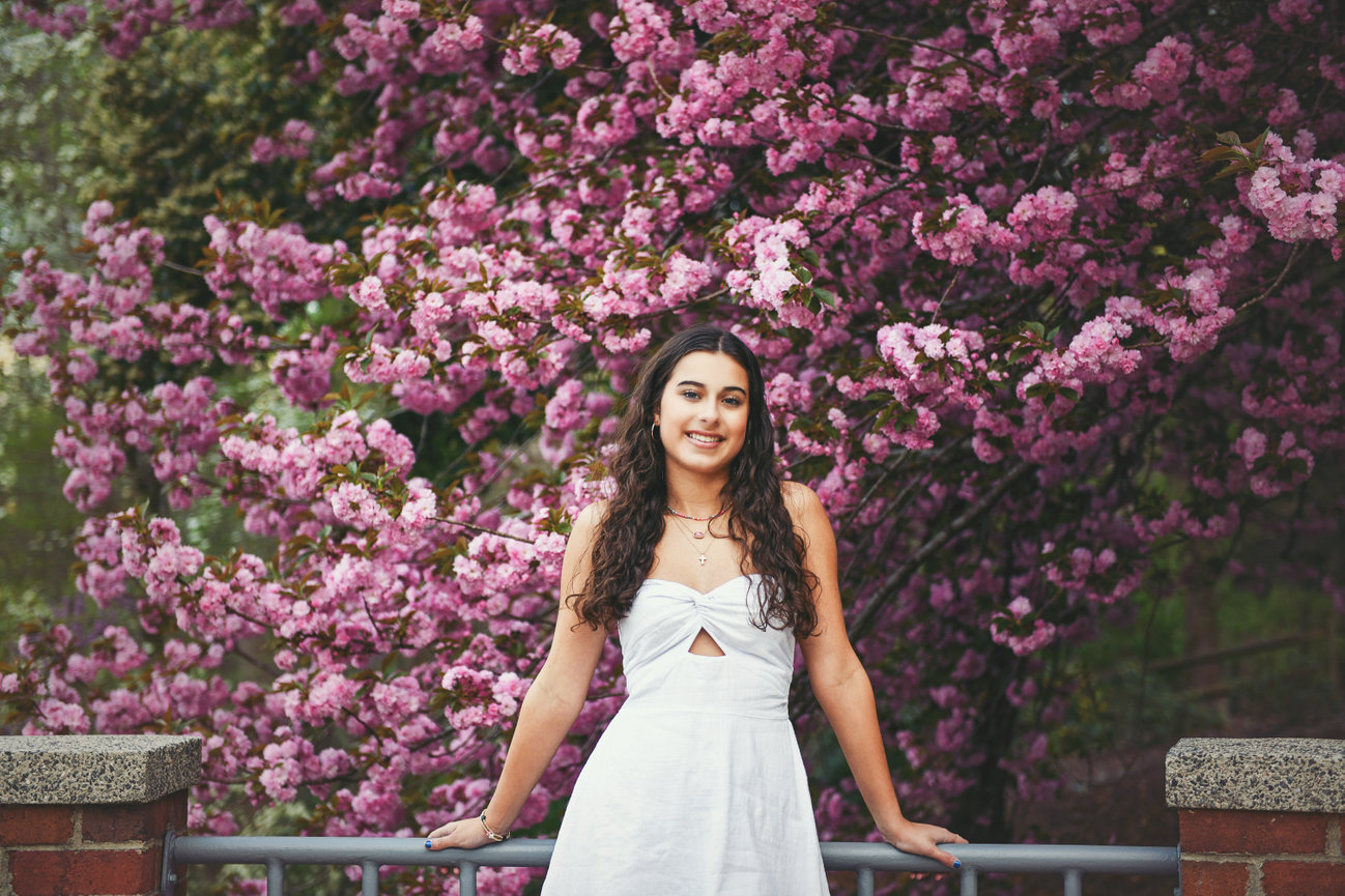 High school senior girl in a white dress standing in front of blooming pink flowers during a spring senior photo session in Clemmons, NC, captured by Megan Gioeli Photography