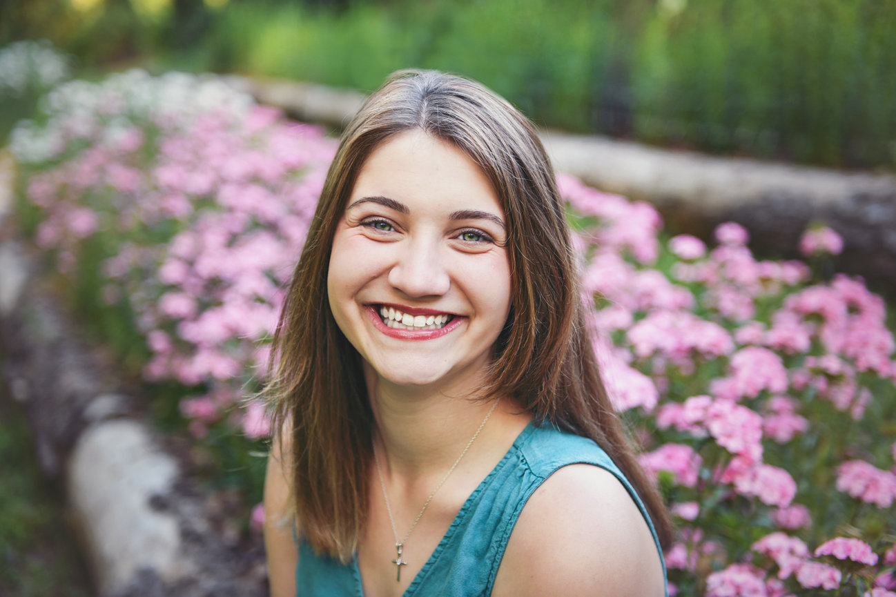 Smiling high school senior girl in a teal shirt sitting in front of blooming pink flowers during a spring photo session in Kernersville, NC at Dewberry Farm, captured by Megan Gioeli Photography