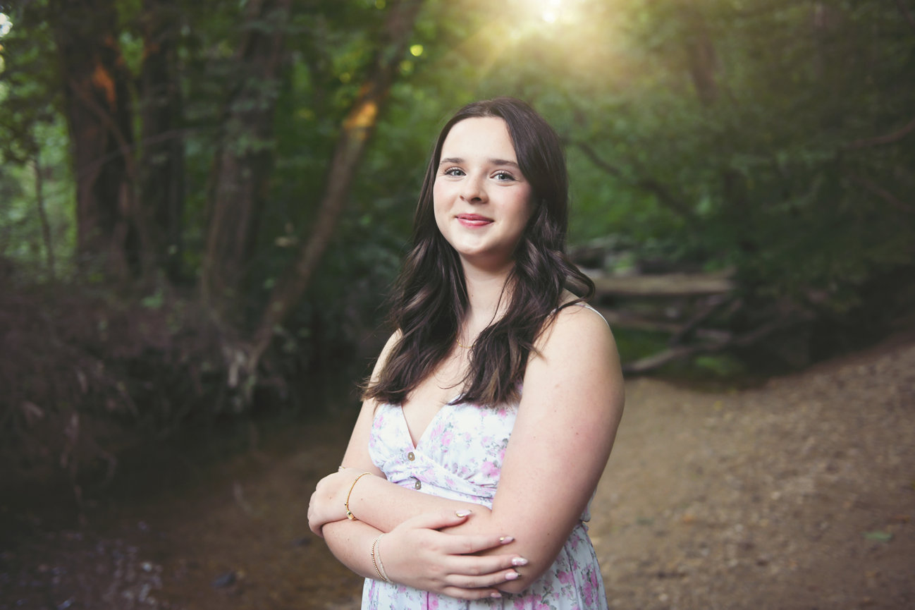 High school senior girl in a floral dress standing confidently in a wooded area with golden sunlight behind her, photographed in Winston-Salem, NC by Megan Gioeli