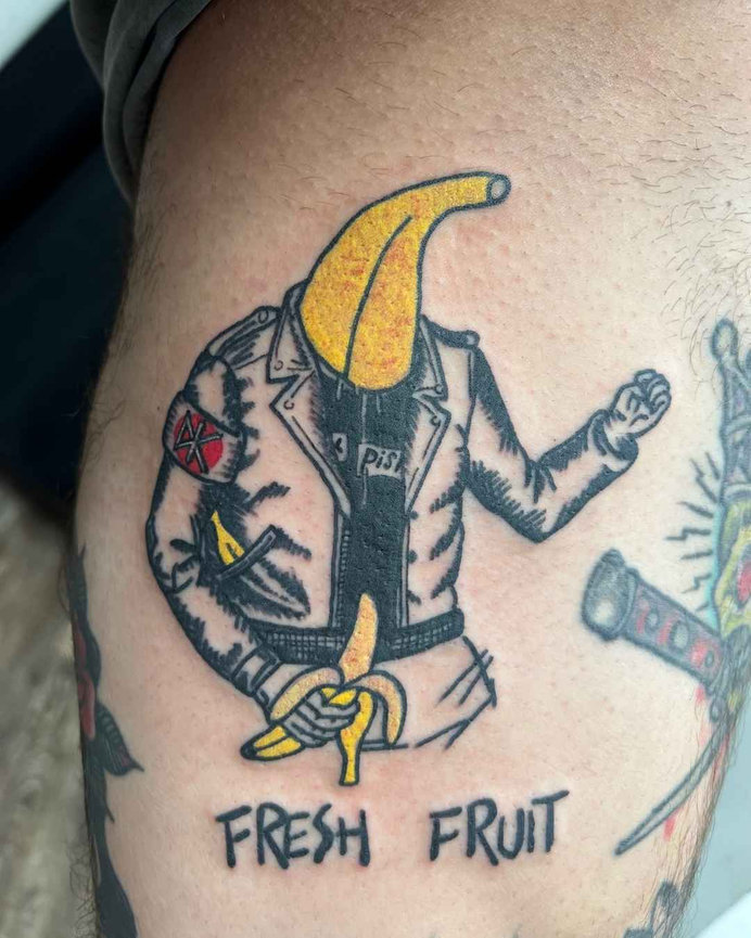 Tattoo by Beam at Torch Tattoo of a banana-headed figure in a leather jacket adorned with punk rock patches, a Sex Pistols t-shirt underneath, and ‘Fresh Fruit’ text underneath.