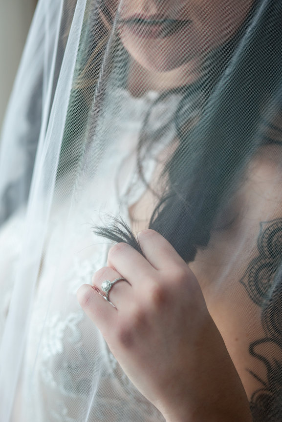 Bride in lace gown holds veil, showing tattoo and gemstone ring.