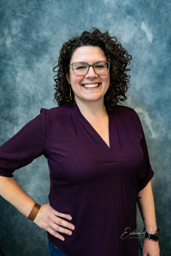 Smiling person with curly hair wearing glasses and a purple top stands against a gray textured background.
