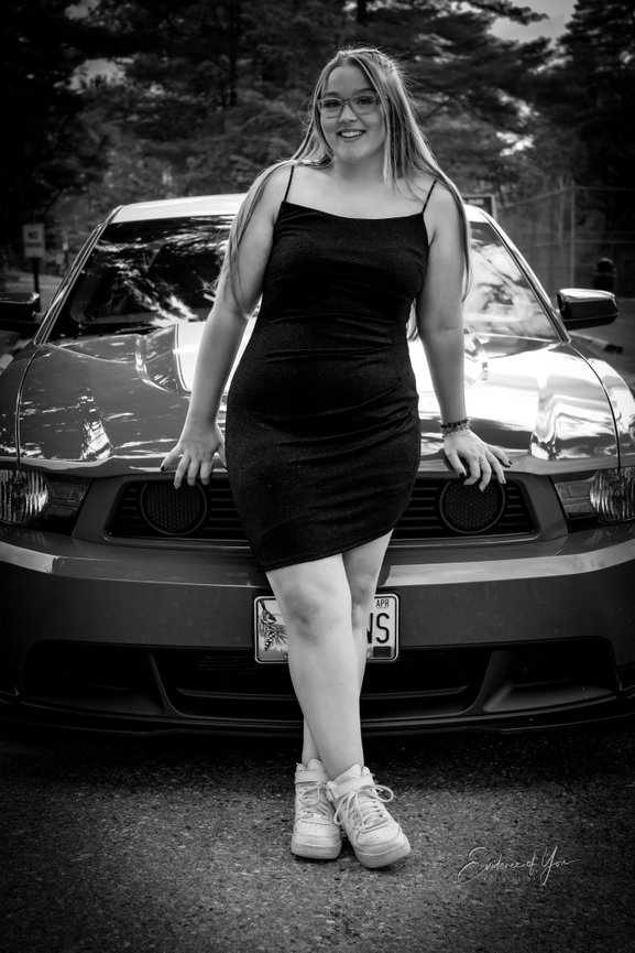 Young woman in a black dress leans against a parked car, smiling. Trees are visible in the background.