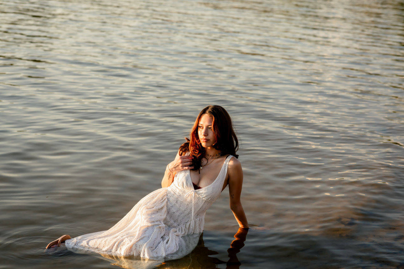 Woman in a white lace dress sitting in the river at sunset during a Texas Hill Country portrait session