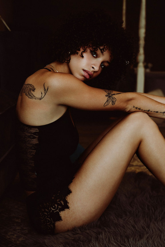 Woman in black lace lingerie posing seated on a fur rug, showing tattoos and curly hair